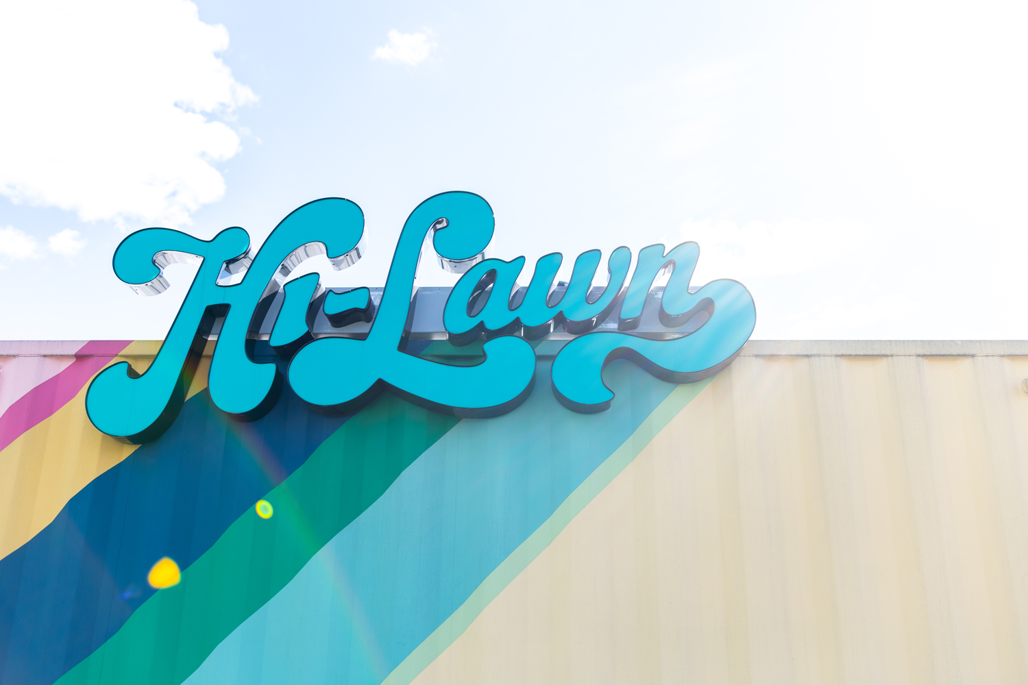 A turquoise blue Hi-Lawn sign in a fun font under a blue sky