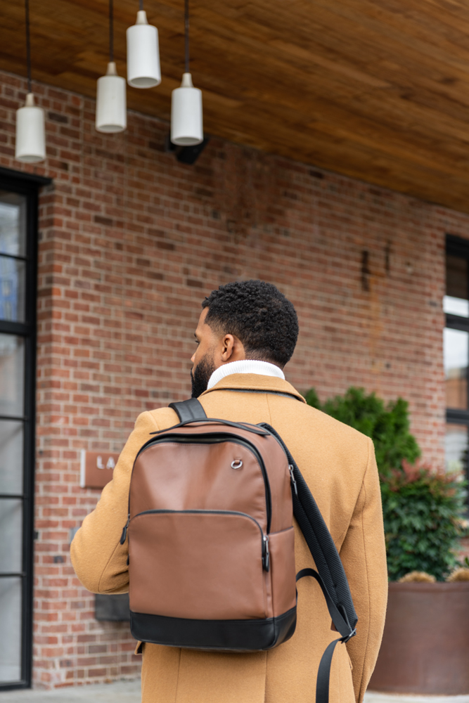 A man in tan overcoat carrying a brown leather backpack outside